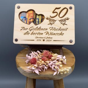May include: A wooden sign with two hearts cut out, a photo frame in the center, and the text "Zur Goldenen Hochzeit die besten W&uuml;nsche Christine & Johann 1974 &hearts; 2024" and the number "50" above the hearts.