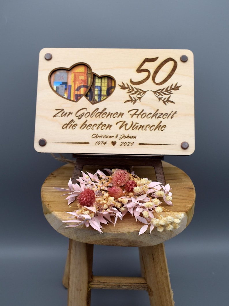 May include: A wooden sign with two hearts cut out, a photo frame in the center, and the text "Zur Goldenen Hochzeit die besten W&uuml;nsche Christine & Johann 1974 &hearts; 2024" and the number "50" above the hearts.