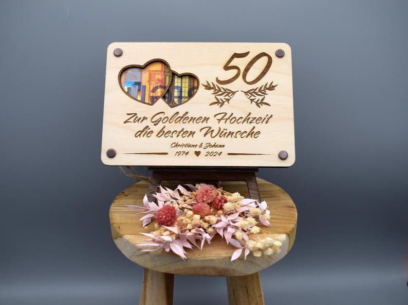 May include: A wooden sign with two heart-shaped cutouts and the number 50 engraved on it. The sign reads "Zur Goldenen Hochzeit die besten W&uuml;nsche Christine & Johann 1974 &hearts; 2024".