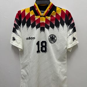 Op de afbeelding: Een wit voetbalshirt met een zwart, rood en geel ruitpatroon op de schouders en kraag. Het shirt toont het nummer 18 in het zwart, het Adidas-logo en het embleem van de Deutscher Fussball-Bund op de voorzijde van de borst.