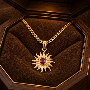 Könnte beinhalten: Eine goldfarbene Halskette mit einem sonnenförmigen Anhänger und einem zentralen roten Edelstein, platziert in einer achteckigen Schatulle aus dunklem Holz. Der Innendeckel enthält ein kleines Licht und ein lasergraviertes Mandala-Motiv.