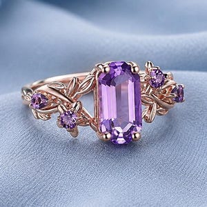 May include: A rose gold ring featuring a central emerald-cut purple amethyst stone flanked by smaller round purple accent stones. The band is designed with intricate metal leaf and vine detailing that extends from the central setting.