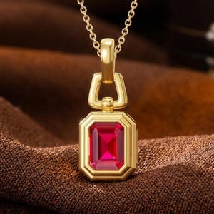 Puede incluir: Un collar con colgante de oro que presenta una piedra preciosa roja vibrante de corte esmeralda rectangular, engastada en un marco de oro pulido. El colgante cuelga de una delicada cadena de oro, con un asa geométrica que une el engaste a la cadena.