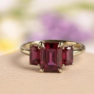 Puede incluir: Un anillo de oro con tres piedras preciosas rectangulares de color rojo intenso. La piedra central es más grande que las dos laterales. El anillo se encuentra sobre un fondo suave y neutro con un fondo floral borroso.