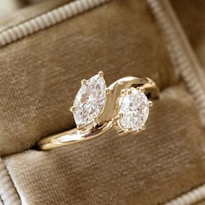 May include: A gold ring with two clear gemstones, one marquise-cut and one round-cut, set in a velvet ring box. The ring box is beige and open, with a second box behind it. The background includes sand and driftwood.