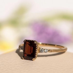 May include: A gold ring with a rectangular, dark red gemstone. The ring has a delicate, ornate design with small accent stones on either side of the main stone. The band is thin and detailed, with a textured pattern.