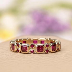 Könnte beinhalten: Ein goldener Ring mit einem sich wiederholenden Muster aus quadratischen und runden roten Edelsteinen. Der Ring liegt auf einer neutralen Oberfläche mit einem verschwommenen lila und grünen Hintergrund.