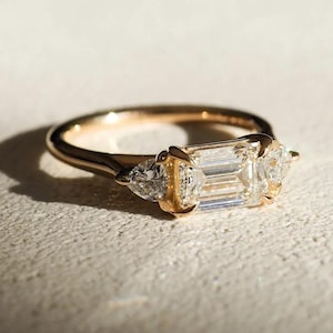 Op de afbeelding: Een gouden ring met een grote rechthoekige diamant in het midden, geflankeerd door twee kleinere driehoekige diamanten. De ring ligt op een lichte ondergrond, met schaduwen.