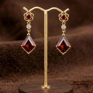 May include: A pair of gold drop earrings featuring a flower-shaped top with a small red stone, a single clear round stone, and a large diamond-shaped red gemstone at the base. The earrings are displayed on a gold metal stand with a round wooden base.