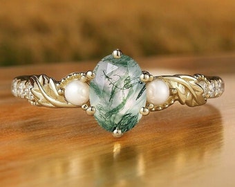 Könnte beinhalten: Ein goldener Ring mit einem ovalen grünen Edelstein, weißen Perlen und blattartigen Details. Der Ring hat kleine klare Steine am Band. Der Hintergrund ist unscharf. Das Design des Rings ist vor einem verschwommenen Hintergrund.