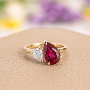 Puede incluir: Un anillo de oro con una piedra preciosa roja oscura en forma de pera y una piedra blanca triangular más pequeña. El anillo tiene una banda de oro pulido y las piedras están juntas.