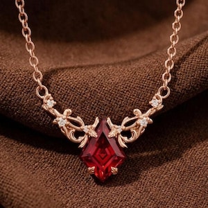 Puede incluir: Un collar de oro rosa con un colgante de piedra preciosa de color rojo intenso en forma de cometa. El engaste presenta un intrincado trabajo de filigrana metálica y pequeñas piedras claras a ambos lados de la gema central, con una cadena fina.