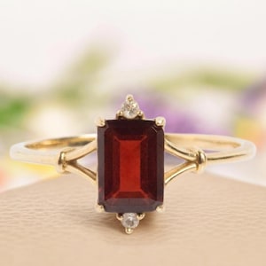 May include: A gold ring with a rectangular, deep red gemstone. The ring has a delicate design with small accent stones at the top and bottom of the rectangular stone. The ring is set against a soft, neutral background.