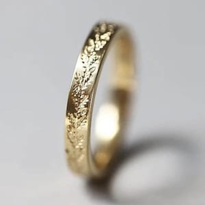 Puede incluir: Un anillo de oro con un detallado patrón floral que rodea la banda. El anillo se encuentra sobre un fondo suave y neutro, resaltando el intrincado diseño. La superficie pulida del anillo refleja la luz, mostrando su delicada artesanía.