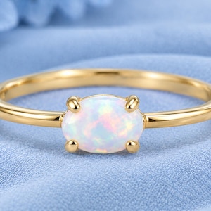 May include: A gold ring featuring a single oval-shaped opal gemstone set in a four-prong setting. The opal displays iridescent flashes of white, pink, and blue light. The band is smooth, polished, and circular, showcasing a simple and clean design.