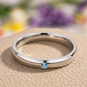 Puede incluir: Un anillo plateado con una peque&ntilde;a gema redonda de color azul claro. El anillo est&aacute; sobre una superficie de tono neutro, con un fondo borroso de flores moradas y amarillas. El anillo tiene un dise&ntilde;o de banda simple.