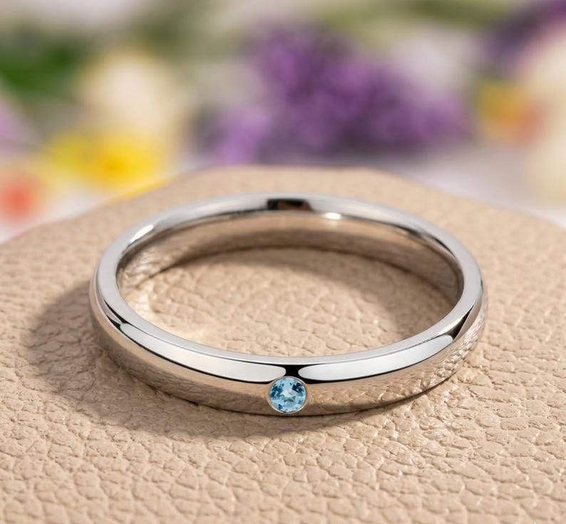 Puede incluir: Un anillo plateado con una peque&ntilde;a gema redonda de color azul claro. El anillo est&aacute; sobre una superficie de tono neutro, con un fondo borroso de flores moradas y amarillas. El anillo tiene un dise&ntilde;o de banda simple.
