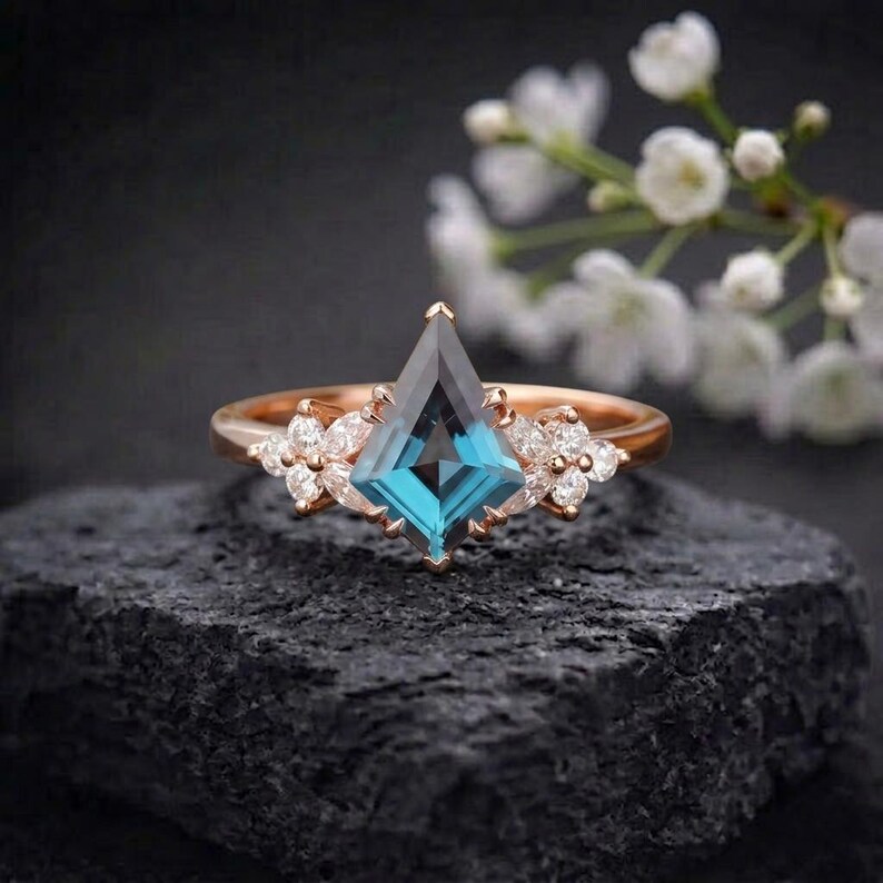 Puede incluir: Anillo de oro rosa con una gema en forma de cometa de color verde azulado. El anillo est&aacute; adornado con peque&ntilde;os grupos de gemas blancas en forma de flor. Se encuentra sobre una superficie oscura y texturizada.