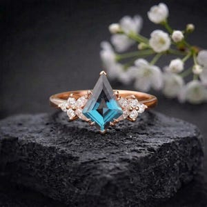 Puede incluir: Un anillo de oro rosa con una gema en forma de cometa de color verde azulado en el centro. El anillo est&aacute; adornado con peque&ntilde;os grupos de gemas blancas en forma de flor a cada lado de la piedra principal. El anillo est&aacute; sobre una superficie oscura y texturizada.