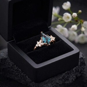 Puede incluir: Un anillo de oro rosa con una gran gema de color verde azulado en forma de cometa y peque&ntilde;os detalles en forma de flor engastados con piedras transparentes. El anillo se muestra en una caja negra.