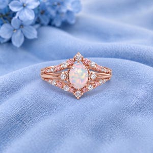 May include: A rose gold ring featuring an oval-shaped opal centre stone. The band is accented with small, round, clear white stones and light pink stones arranged in a symmetrical, pointed design that extends outward from the central opal.