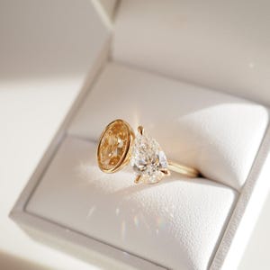 Op de afbeelding: Een gouden ring met twee edelstenen, een ovale, goudkleurige steen en een heldere, peervormige diamant. De ring wordt gepresenteerd in een witte doos. De band is slank en goud. Een elegante ring.