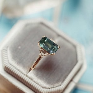 Può includere: Un anello d'oro con una grande pietra preziosa color verde acqua, taglio smeraldo. L'anello ha una fascia sottile ed è incastonato con pietre più piccole. Presentato in un portagioie di velluto grigio, su uno sfondo blu tenue.
