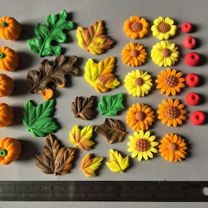 May include: A collection of small, moulded autumn-themed decorations including orange pumpkins, green and brown oak leaves, yellow and orange maple leaves, yellow sunflowers, and red berries, arranged above a metal ruler measuring 15 centimetres.