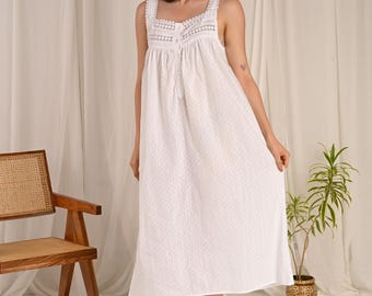 May include: A white, sleeveless nightgown with a square neckline and delicate lace trim. The dress features a button-down front and a subtle, all-over patterned design. The model is barefoot and standing in front of a white curtain.