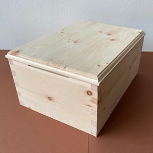 Könnte beinhalten: Eine natürliche Holzkiste mit einem Scharnierdeckel und einem einfachen Design. Die Kiste ist aus hellem Holz mit einer natürlichen Oberfläche.