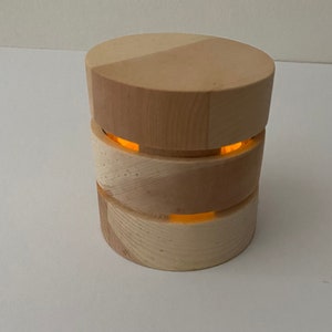 Peut inclure: Une lampe cylindrique en bois avec une lueur orange chaude émanant des espaces entre les sections circulaires empilées. La lampe est faite de bois de couleur claire, avec un motif de grain naturel visible sur la surface.