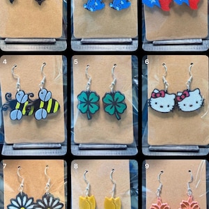 May include: A collection of nine pairs of dangle earrings displayed on individual brown cards, featuring various designs including orcas, blue characters, the Texas flag, bees, four-leaf clovers, white cat faces, daisies, yellow tulips, and pink flowers.