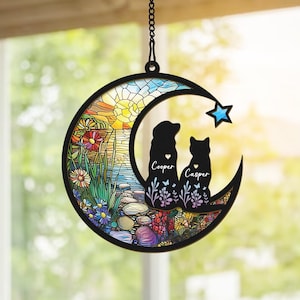 May include: A stained glass moon-shaped suncatcher with a dog and cat silhouette. The suncatcher features a colorful stained glass design with a sunset scene and flowers. The dog and cat are facing the sunset and have the names "Cooper" and "Casper" written below them.