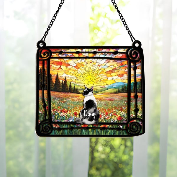 May include: A stained glass window panel featuring a black and white cat named Daisy, set against a colorful landscape with a sunset. The panel is framed in black and hangs from a black chain.