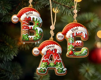 May include: Three hanging Christmas ornaments shaped like gingerbread letters B, A, and E. Each letter features a red Santa hat, white icing, green and red patterns, and the year 2026. The names Bella, Annie, and Elise are printed on the hats.
