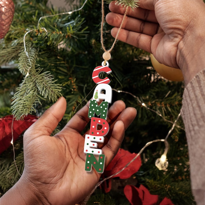 May include: A Christmas ornament with the name "CAMDEN" spelled out in vertical letters. Each letter is decorated with a different Christmas pattern, including red and white stripes, green and white polka dots, and a small Christmas tree.