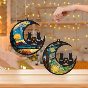 May include: Two wooden moon-shaped ornaments with a starry night painting. Each ornament features two cats sitting back to back with a heart between them. The names "Anna & David" and "Emma & Peter" are written on the ornaments.