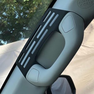 Puede incluir: Accesorio de coche de plástico negro con un mango gris y un patrón de rejilla. El accesorio está unido al parabrisas de un coche.