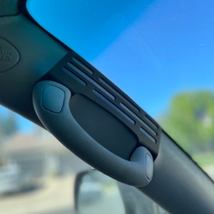 Op de afbeelding: Een zwarte auto-interieur handgreep met een textuur en een zwarte plastic ventilatiekap met een reeks horizontale sleuven.