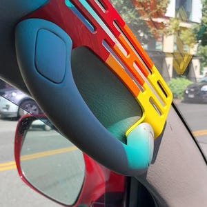 May include: A gray car grab handle featuring a decorative, flat, rectangular attachment with a gradient color scheme transitioning from red at the top to orange in the middle and yellow at the bottom, with three horizontal cutouts.