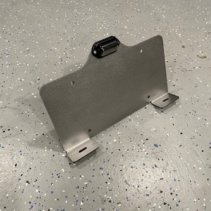 May include: A gray metal license plate holder with a black light fixture attached to the top. The holder has two mounting brackets on the bottom.