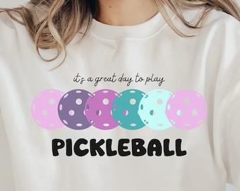 May include: White sweatshirt with the text "it's a great day to play pickleball" and a row of colorful pickleball paddles.