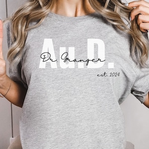 May include: A gray t-shirt with the text "Au.D. Dr. Granger est. 2024" printed on the front.