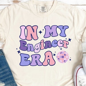 May include: A light beige t-shirt with the text "IN MY Engineer ERA" in pink, purple, and blue letters with a disco ball graphic. The shirt is on a wooden surface with a pair of white sneakers and a pair of blue denim shorts.