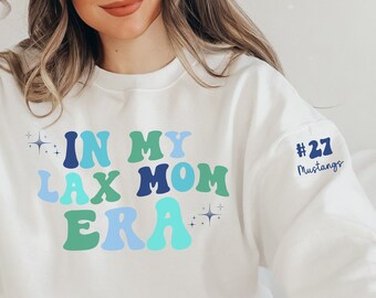 May include: White sweatshirt with the text "In My Lax Mom Era" in colorful retro font with sparkling stars. The right sleeve has the text "#27 Mustangs" in blue.