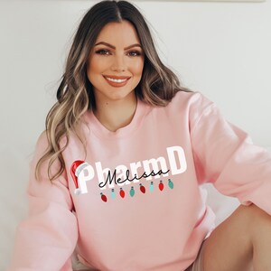 May include: Pink sweatshirt with the text "PharmD Melissa" in white with a red Santa hat and a string of colorful Christmas lights.