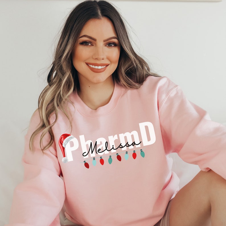 May include: Pink sweatshirt with the text "PharmD Melissa" in white with a red Santa hat and a string of colorful Christmas lights.