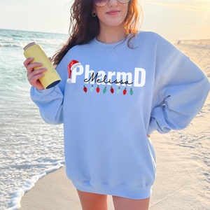 May include: Light blue sweatshirt with a white graphic of a Santa hat, the text "PharmD" and the name "Melissa" written in cursive. The graphic also includes a string of colorful Christmas lights.