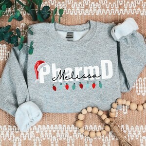 May include: A gray sweatshirt with the text "PharmD Melissa" in white with a red Santa hat and a string of colorful lights.