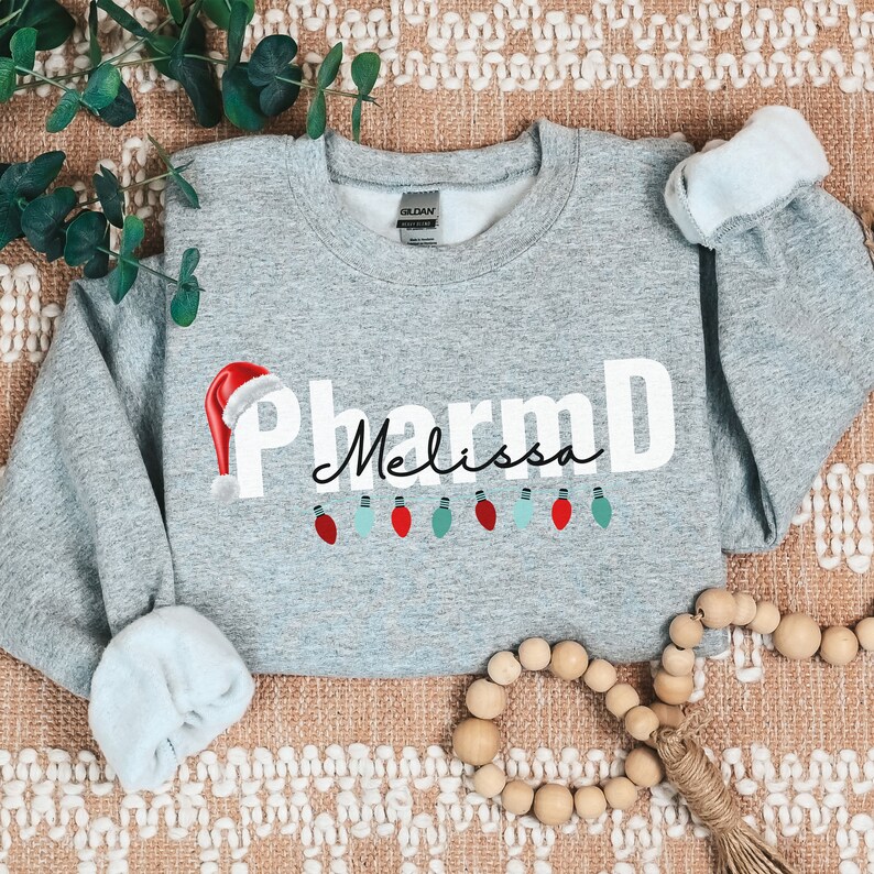 May include: A gray sweatshirt with the text "PharmD Melissa" in white with a red Santa hat and a string of colorful lights.